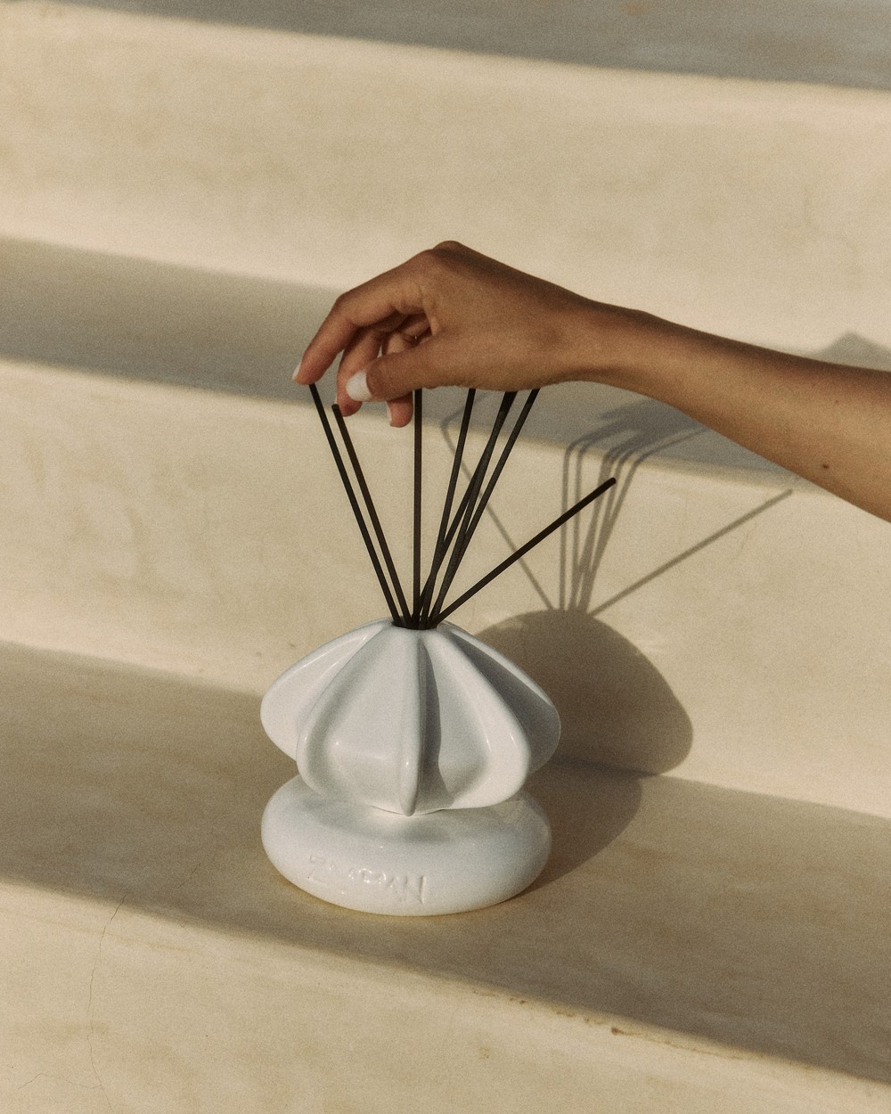 Hand fanning reed diffuser sticks in white Tourniquet ceramic vessel, sunlit steps